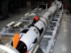 Long white and black rocket mounted on a metal test cradle in a laboratory, with technicians nearby and orange safety fins visible.