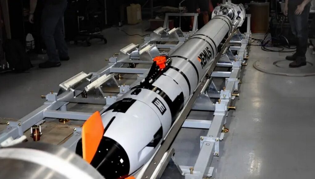 Long white and black rocket mounted on a metal test cradle in a laboratory, with technicians nearby and orange safety fins visible.