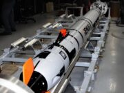 Long white and black rocket mounted on a metal test cradle in a laboratory, with technicians nearby and orange safety fins visible.
