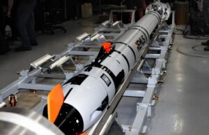 Long white and black rocket mounted on a metal test cradle in a laboratory, with technicians nearby and orange safety fins visible.