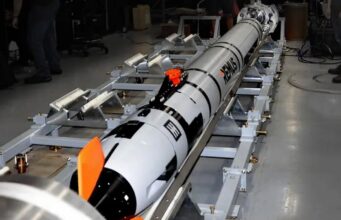 Long white and black rocket mounted on a metal test cradle in a laboratory, with technicians nearby and orange safety fins visible.