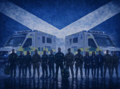 Emergency responders in uniform stand in front of two NHS emergency ambulances with a Scottish flag backdrop behind them.