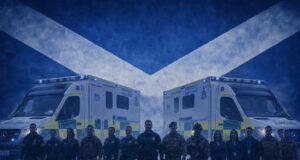 Emergency responders in uniform stand in front of two NHS emergency ambulances with a Scottish flag backdrop behind them.