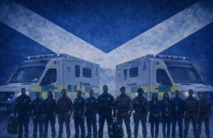 Emergency responders in uniform stand in front of two NHS emergency ambulances with a Scottish flag backdrop behind them.
