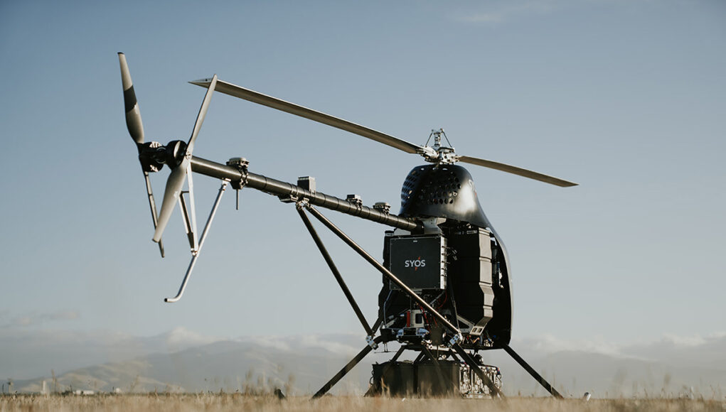 syos-sa200-box A small experimental helicopter drone with a dual-rotor tail and landing skids sits in a field under a clear sky.