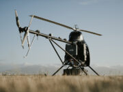 A small experimental helicopter drone with a dual-rotor tail and landing skids sits in a field under a clear sky.