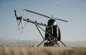 SYOS autonomous heavy-lift helicopter clears serial production A small experimental helicopter drone with a dual-rotor tail and landing skids sits in a field under a clear sky.