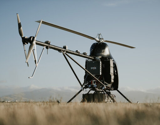 A small experimental helicopter drone with a dual-rotor tail and landing skids sits in a field under a clear sky.