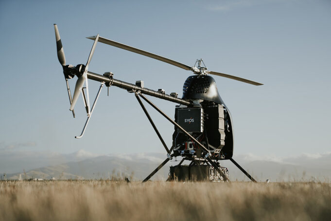 SYOS autonomous heavy-lift helicopter clears serial production A small experimental helicopter drone with a dual-rotor tail and landing skids sits in a field under a clear sky.