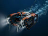 SYOS unveils underwater drone to complete multi-domain portfolio Underwater orange-and-black submersible drone with lights cruising through deep blue water.
