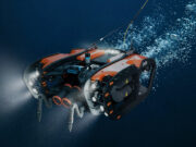 SYOS unveils underwater drone to complete multi-domain portfolio Underwater orange-and-black submersible drone with lights cruising through deep blue water.