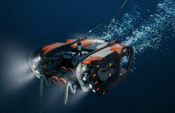 SYOS unveils underwater drone to complete multi-domain portfolio Underwater orange-and-black submersible drone with lights cruising through deep blue water.