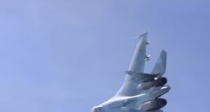 Blurred fighter jet ascending into a clear blue sky, twin vertical tails visible at the rear.
