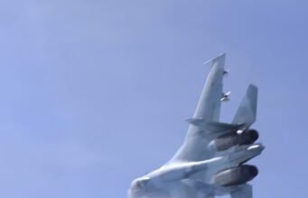 Blurred fighter jet ascending into a clear blue sky, twin vertical tails visible at the rear.