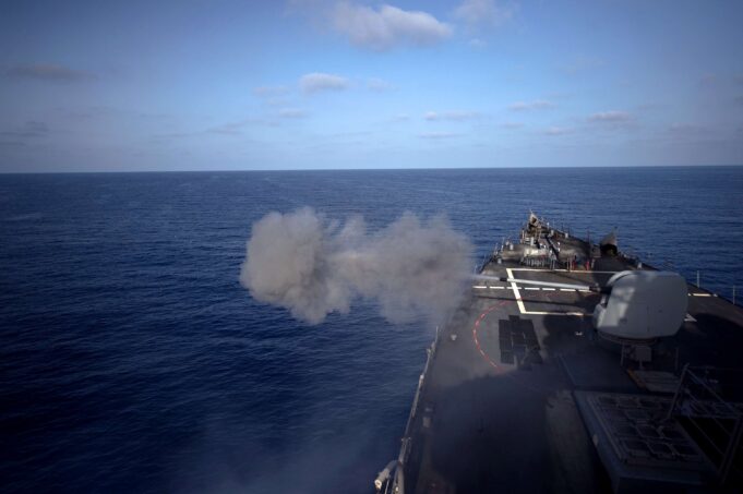 Military ship&rsquo;s flight/deck at sea with a smoke plume near the bow and open ocean ahead.
