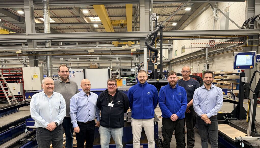 Eight people pose for a group photo in a manufacturing plant, standing in front of large automated equipment and conveyors.