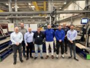 Eight people pose for a group photo in a manufacturing plant, standing in front of large automated equipment and conveyors.