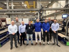 Eight people pose for a group photo in a manufacturing plant, standing in front of large automated equipment and conveyors.