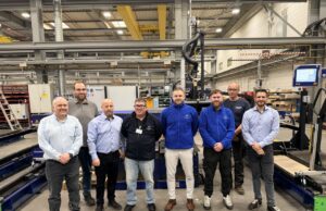 Eight people pose for a group photo in a manufacturing plant, standing in front of large automated equipment and conveyors.