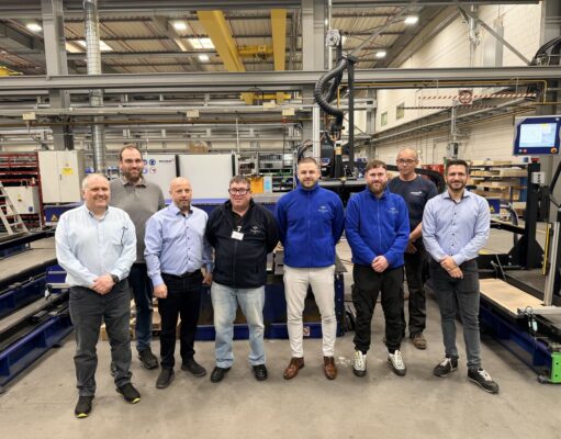 Eight people pose for a group photo in a manufacturing plant, standing in front of large automated equipment and conveyors.