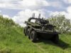 Eight-wheeled armored military vehicle on a grassy slope under a cloudy sky, with equipment racks and antennas mounted on top.