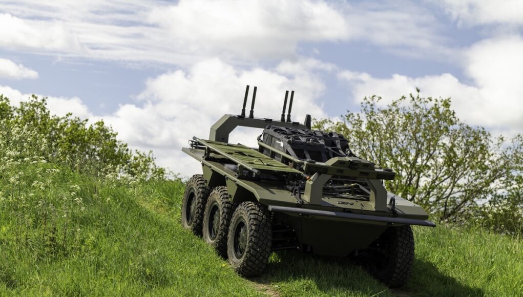 Eight-wheeled armored military vehicle on a grassy slope under a cloudy sky, with equipment racks and antennas mounted on top.