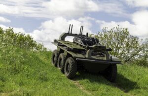 Eight-wheeled armored military vehicle on a grassy slope under a cloudy sky, with equipment racks and antennas mounted on top.