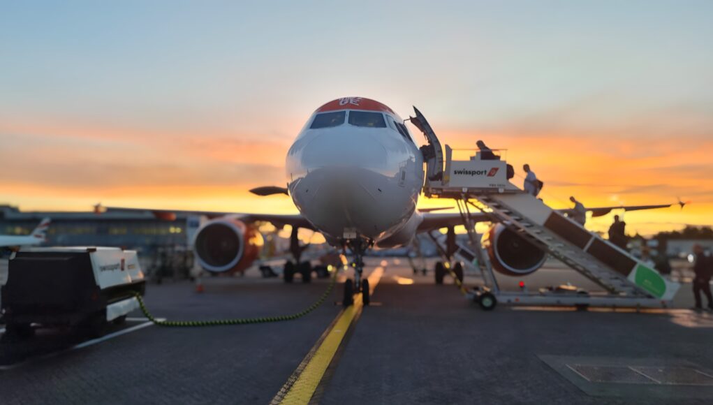 20250909_063320.remini-enhanced Front view of a passenger airliner at the gate with a jet bridge and sunset in the background, boarding area visible behind it.