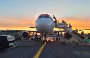 Front view of a passenger airliner at the gate with a jet bridge and sunset in the background, boarding area visible behind it.