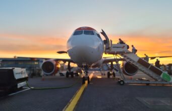 Front view of a passenger airliner at the gate with a jet bridge and sunset in the background, boarding area visible behind it.