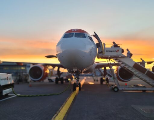 Front view of a passenger airliner at the gate with a jet bridge and sunset in the background, boarding area visible behind it.