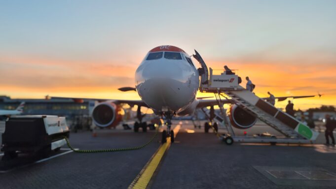 Front view of a passenger airliner at the gate with a jet bridge and sunset in the background, boarding area visible behind it.