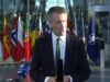 Frozen Russian assets to fund UK drones for Ukraine Male news reporter in a dark suit and green tie speaks into a handheld microphone outside a glass-fronted building, with a row of international flags behind him.