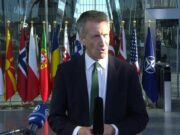 Male news reporter in a dark suit and green tie speaks into a handheld microphone outside a glass-fronted building, with a row of international flags behind him.