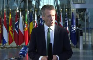 Frozen Russian assets to fund UK drones for Ukraine Male news reporter in a dark suit and green tie speaks into a handheld microphone outside a glass-fronted building, with a row of international flags behind him.