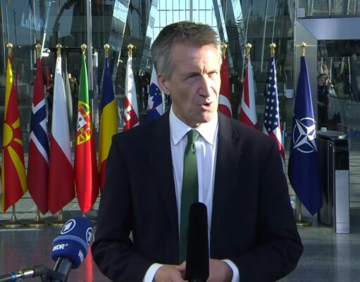 Male news reporter in a dark suit and green tie speaks into a handheld microphone outside a glass-fronted building, with a row of international flags behind him.