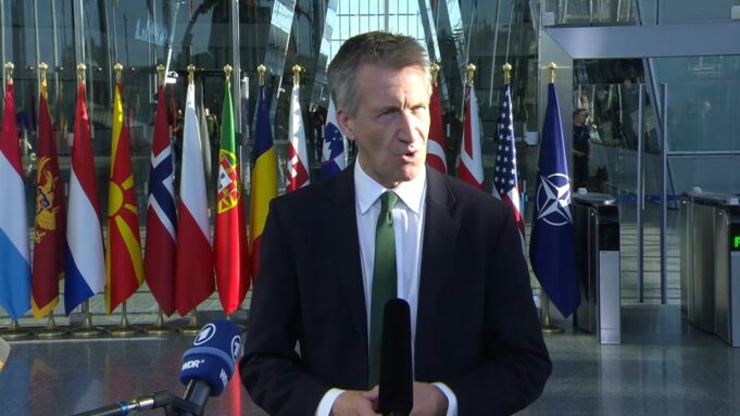 Frozen Russian assets to fund UK drones for Ukraine Male news reporter in a dark suit and green tie speaks into a handheld microphone outside a glass-fronted building, with a row of international flags behind him.