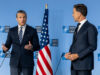 Hegseth presses NATO allies as US ramps up spending Two men in dark suits speak at a NATO press conference with the American flag between them.