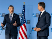 Two men in dark suits speak at a NATO press conference with the American flag between them.
