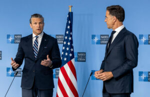 Hegseth presses NATO allies as US ramps up spending Two men in dark suits speak at a NATO press conference with the American flag between them.