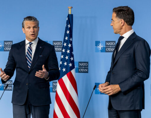 Two men in dark suits speak at a NATO press conference with the American flag between them.