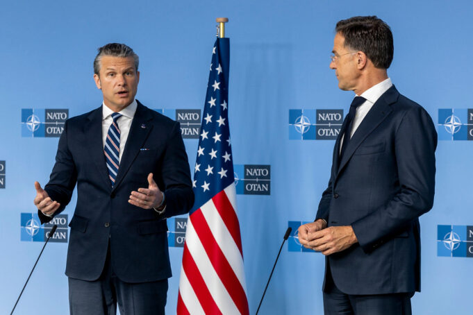 Hegseth presses NATO allies as US ramps up spending Two men in dark suits speak at a NATO press conference with the American flag between them.