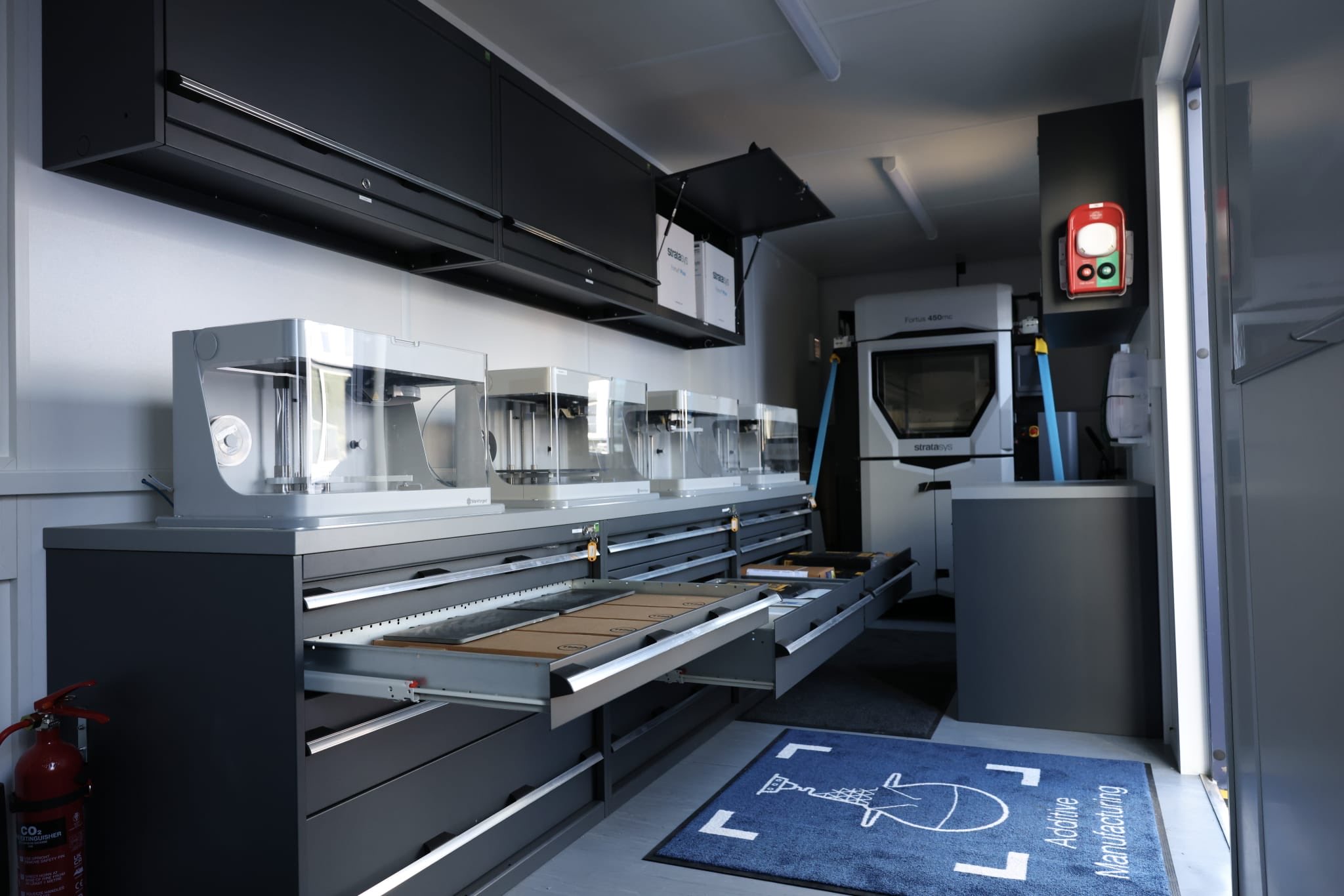 Interior of a mobile lab with black overhead cabinets, a Fortus 450mc 3D printer at the back, and clear plastic enclosures on a workbench toward the left.