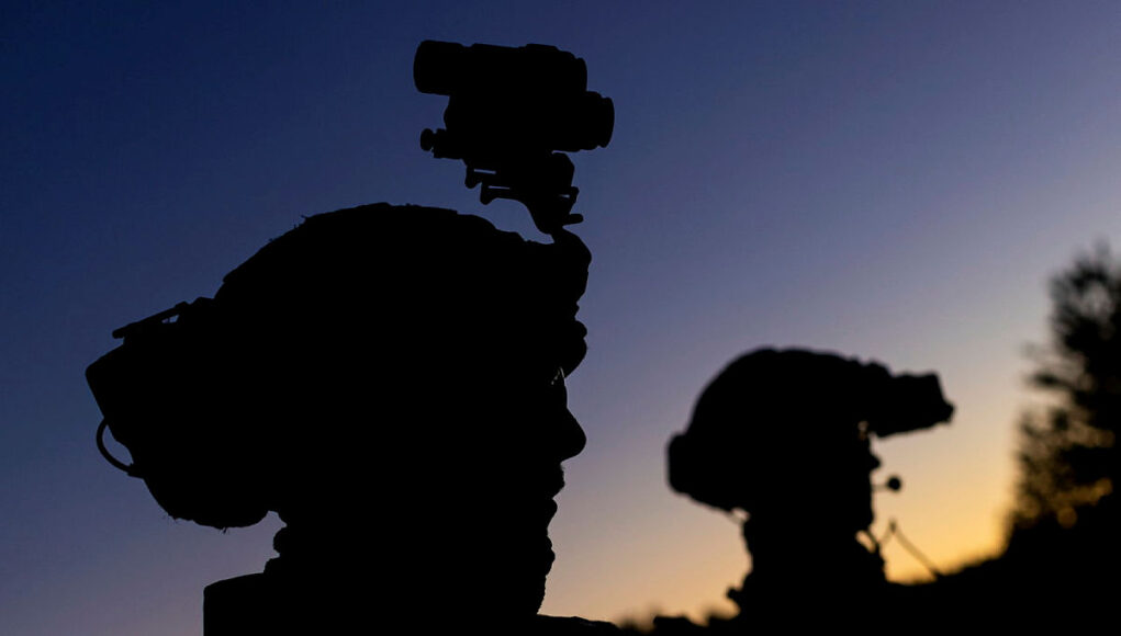 Silhouetted soldiers with gear and rifles against a dusky sunset, one with a helmet-mounted device.