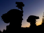 Silhouetted soldiers with gear and rifles against a dusky sunset, one with a helmet-mounted device.