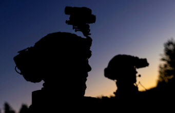 Silhouetted soldiers with gear and rifles against a dusky sunset, one with a helmet-mounted device.