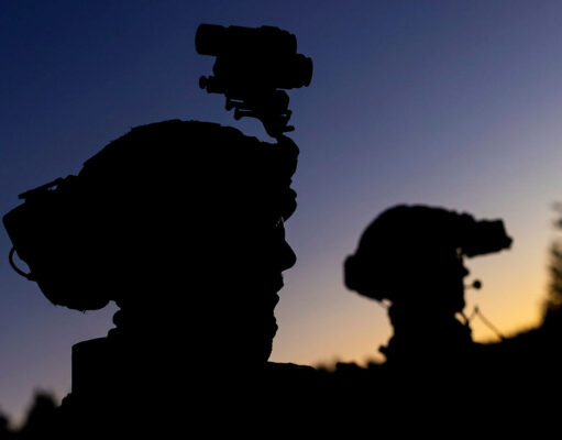 Silhouetted soldiers with gear and rifles against a dusky sunset, one with a helmet-mounted device.