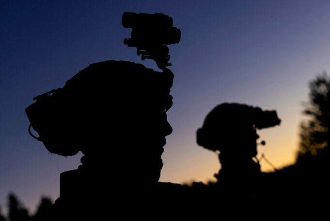 Silhouetted soldiers with gear and rifles against a dusky sunset, one with a helmet-mounted device.