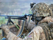 Soldier in desert camouflage gear and helmet feeding a belt-fed machine gun in tall grass.