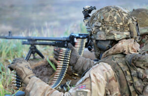 Heckler & Koch set to supply UK machine guns for a decade Soldier in desert camouflage gear and helmet feeding a belt-fed machine gun in tall grass.